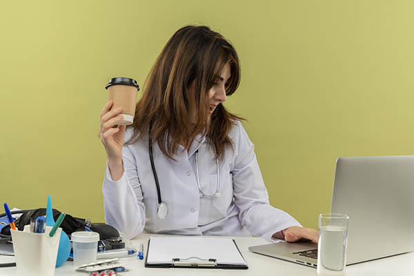 Impressionada médica de meia-idade com túnica médica e estetoscópio sentada à secretária com ferramentas médicas e prancheta usando um portátil segurando um copo de café de plástico isolado sobre fundo verde azeitona